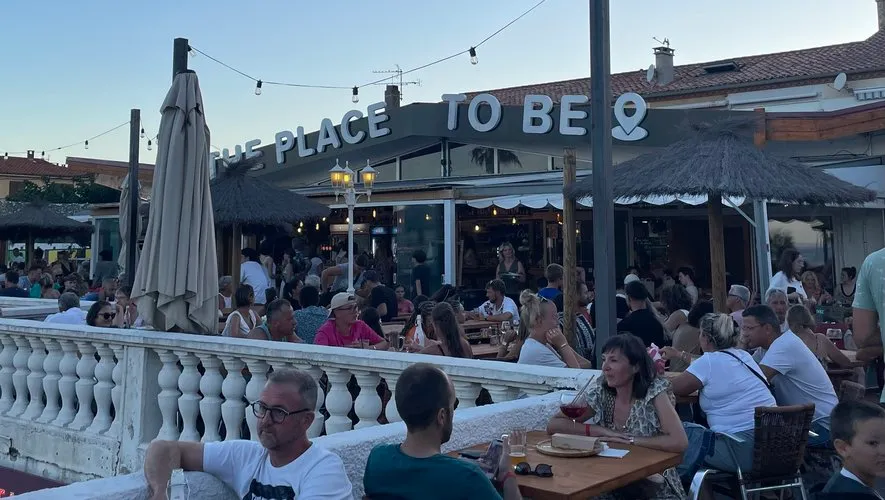 Terrasse animée de The Place to Be en soirée, enseigne lumineuse et kiosques en arrière-plan